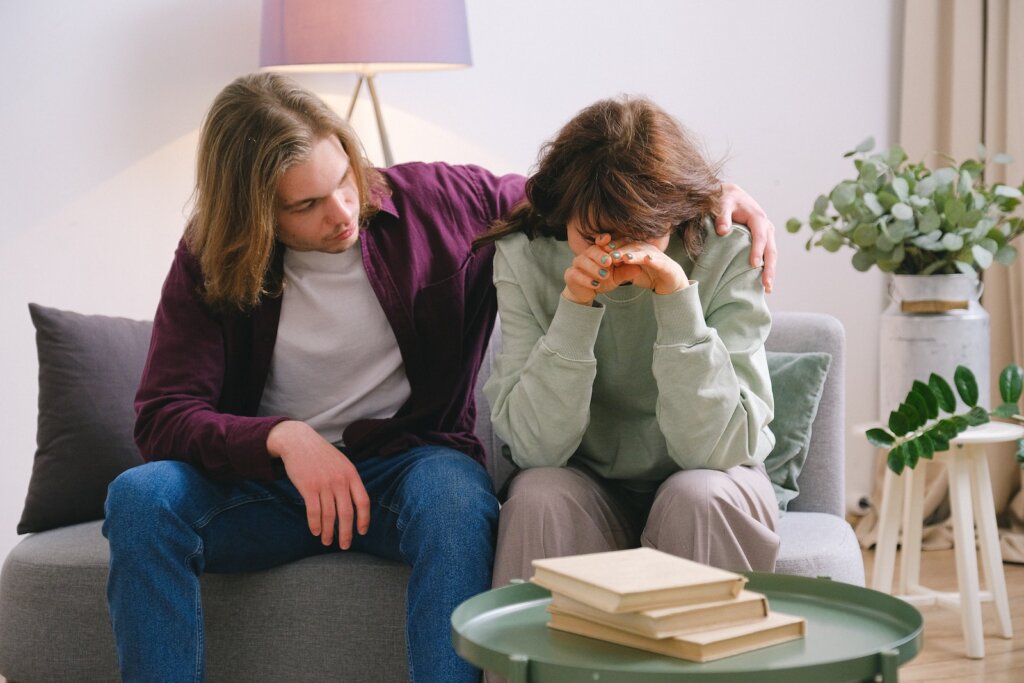 Young male in jeans comforting depressed upset female crying on gray sofa near table with books